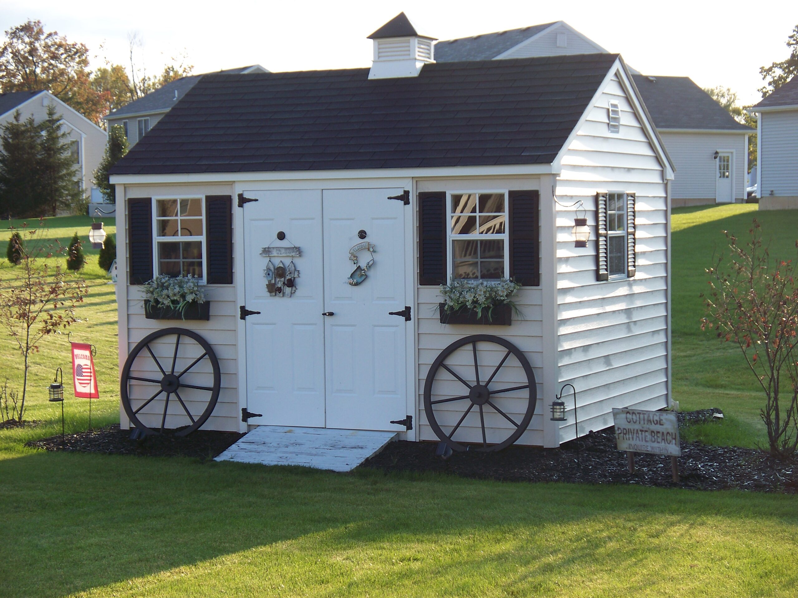 A white cottage shed with blue trim and roofing, decorated with decorative wooden wheels against the wall, hanging lanterns, and door-hangers.